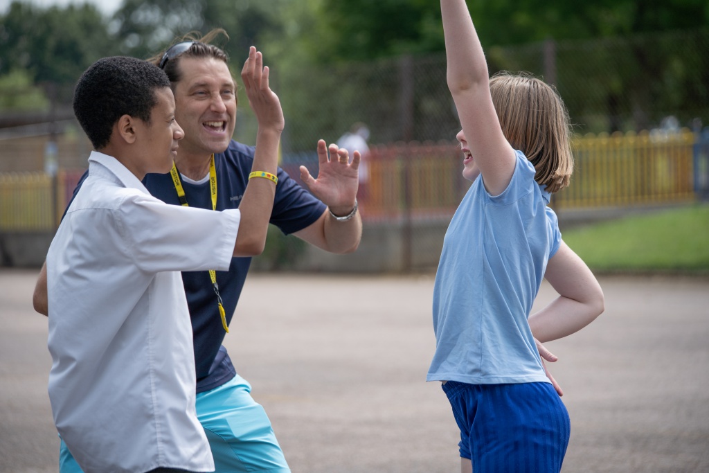 A male PE teacher is pictured smiling outdoors, alongside two students, a boy and a girl.