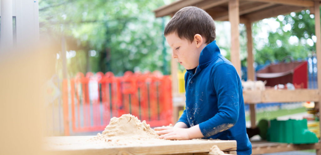 A young boy can be seen playing with some sand on a wooden table, outdoors on the academy grounds.