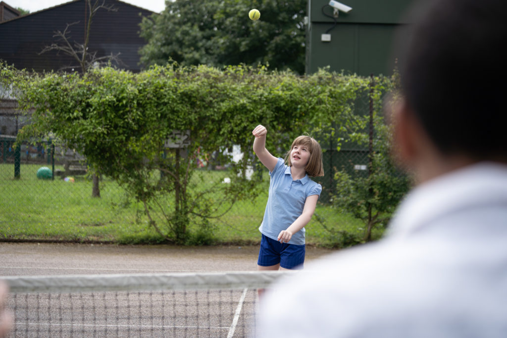 A girl in PE uniform throwing a tennis ball over the net on a tennis court