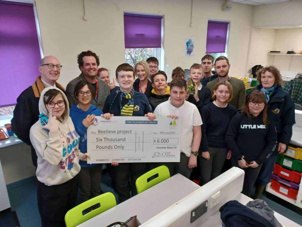 Students are seen welcoming visitors to the academy from the Beelieve Project. Three students can be seen holding a large cheque with a charity donation written on.