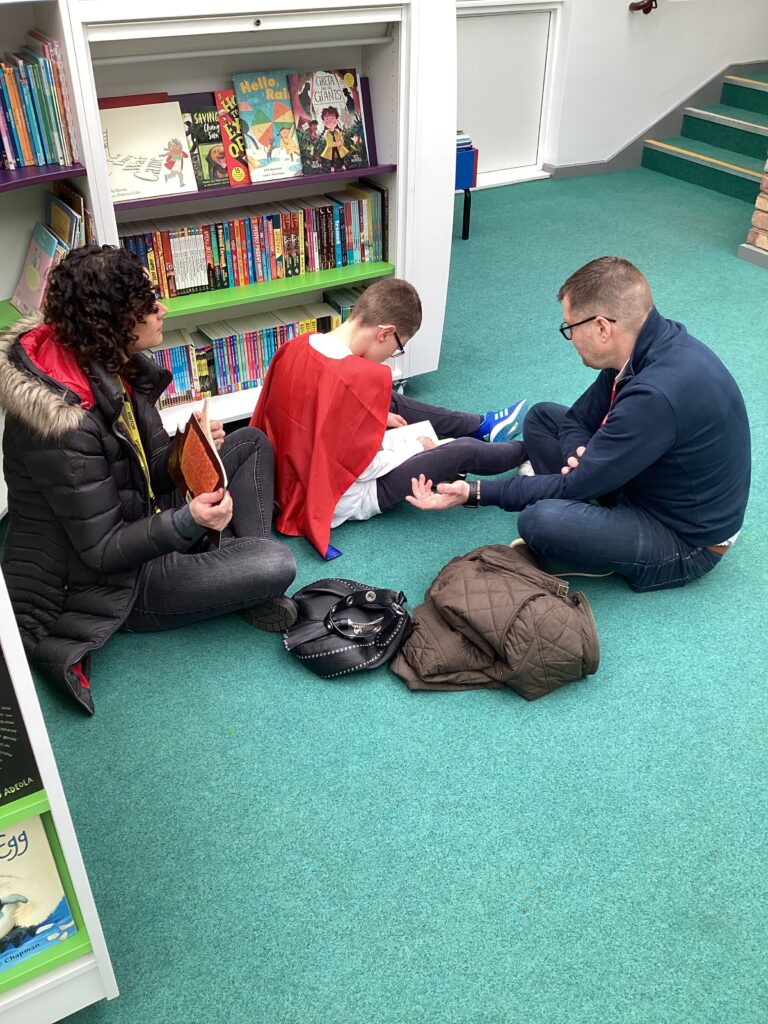 A boy is shown sitting on the floor, reading a book with two adults on World Book Day 2023.