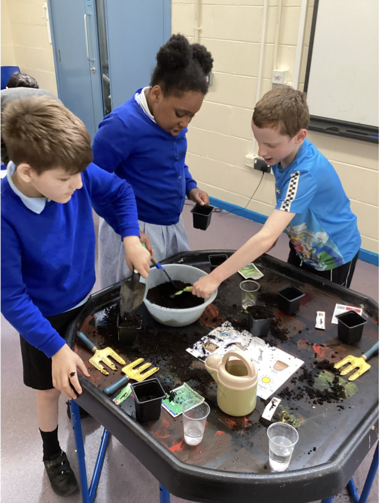 Three students, two boys and a girl, are pictured using soil to fill some plant pots on a table in a corridor of the academy building.