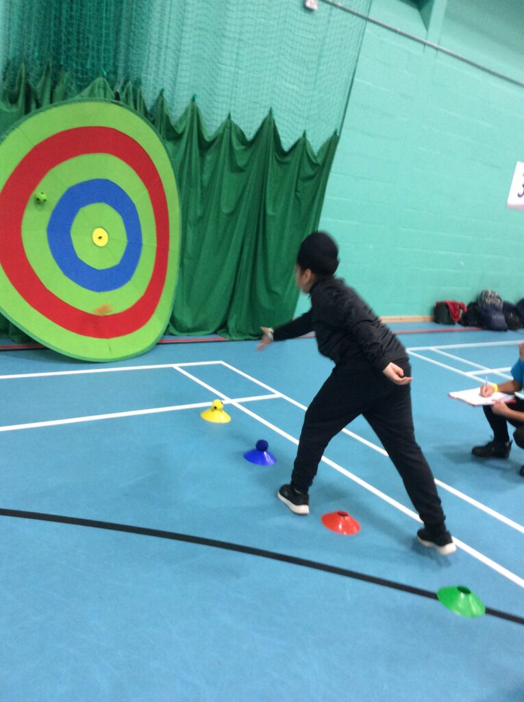 A young, male student can be seen having just thrown a dart at a large plastic target in the Sports Hall, as part of a school-wide competition.