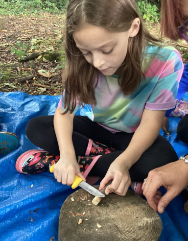 A female student is pictured wearing her own clothes outdoors in the forest, using a knife to shave the bark off of a tree branch,