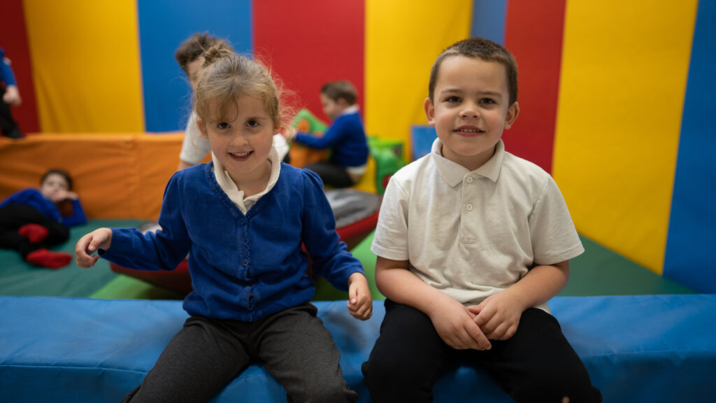 2 young children sitting in a soft play area smiling for the photo