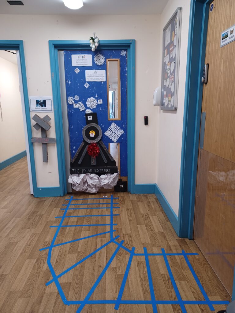 A Phase 4 classroom door is seen decorated for Christmas with an image of a train on the front. Train tracks are seen stuck to the floor in front of the door.