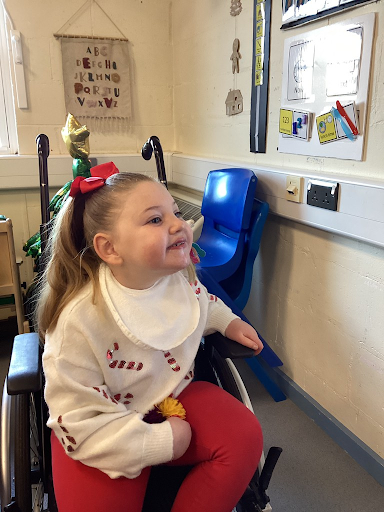 A young girl is seen sat down in her wheelchair, smiling whilst wearing her Christmas Jumper for Christmas Jumper Day 2023.