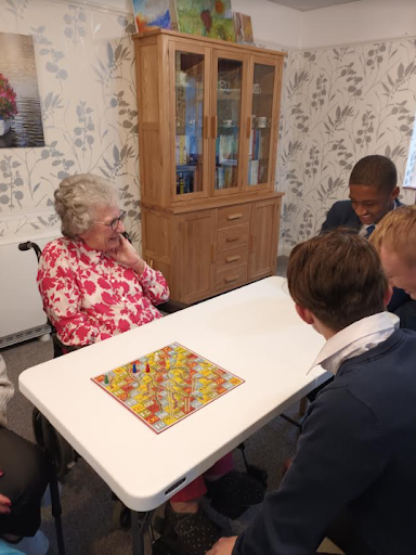 An elderly resident at a retirement home is seen playing a board game with some Milestone Academy students and smiling.
