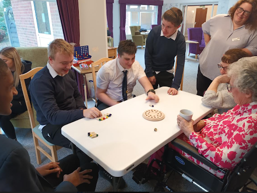 An elderly resident at a retirement home is seen playing a board game with some Milestone Academy students and smiling.