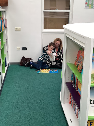 A student is pictured sat on the floor in a Library area, alongside his mother, reading a book together during a Reading Brunch session.