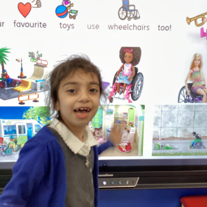 A young girl is pictured smiling for the camera, whilst pointing at some pictures on an Interactive Board.