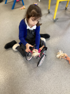 A young girl is seen playing with a doll by putting it into a wheelchair and moving it around on the carpet.