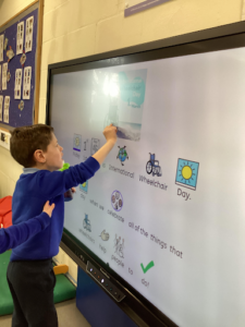 A young boy is seen drawing something on the Interactive Board at the front of the classroom.