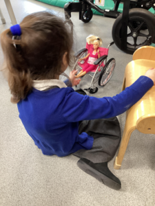 A young girl is shown playing with a Barbie doll by sitting it in a wheelchair and moving it about on the carpet.