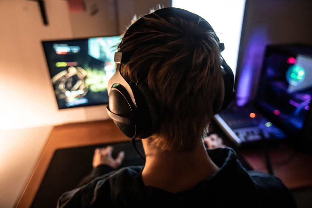 A young man seen from behind, sitting at his desk wearing headphones and playing a game on his PC.